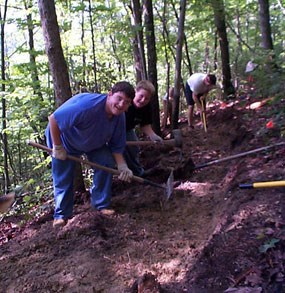 Two boys using hand tools pose for the camera as they dig a trench for a trail