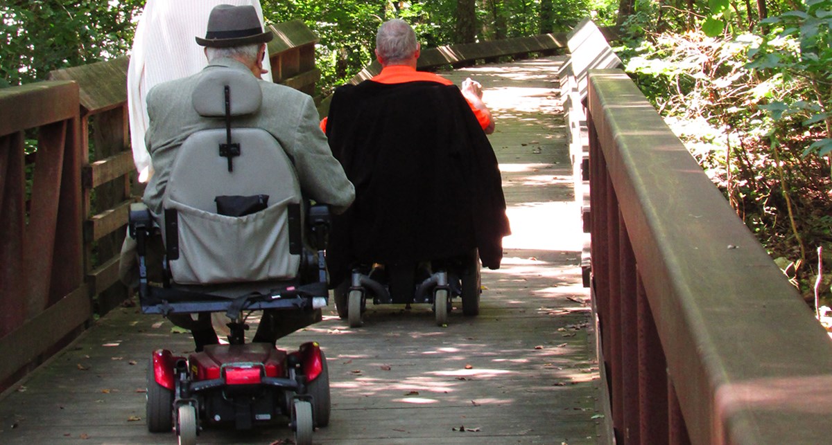 Two motorized wheelchair users on a wooden boardwalk.