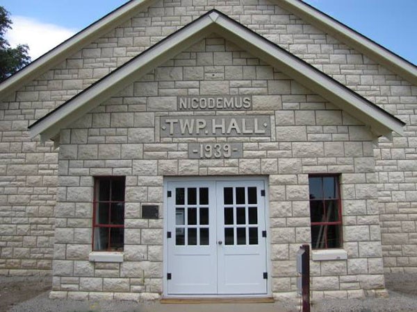 The front of a limestone building with white double doors. There is one window on each side of the doors. Engraved in the limestone above the door is three rows of text that reads "NICODEMUS, TWP HALL, 1939"