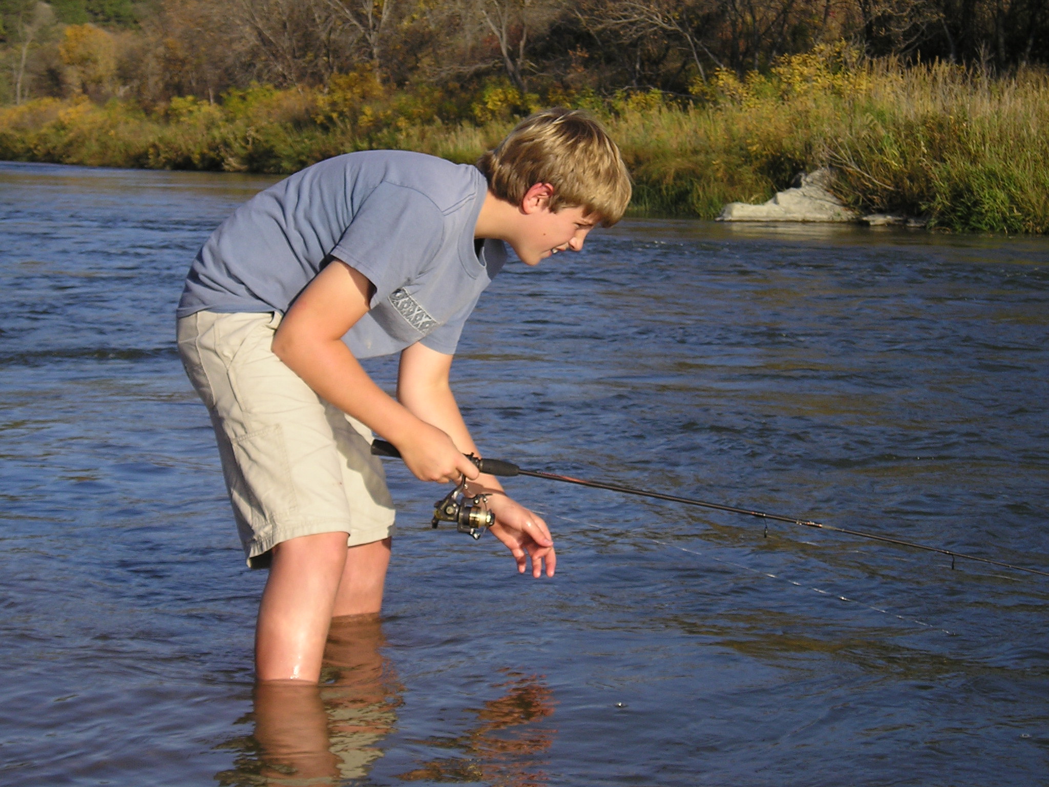 A young man bends over running water with a fishing pole.