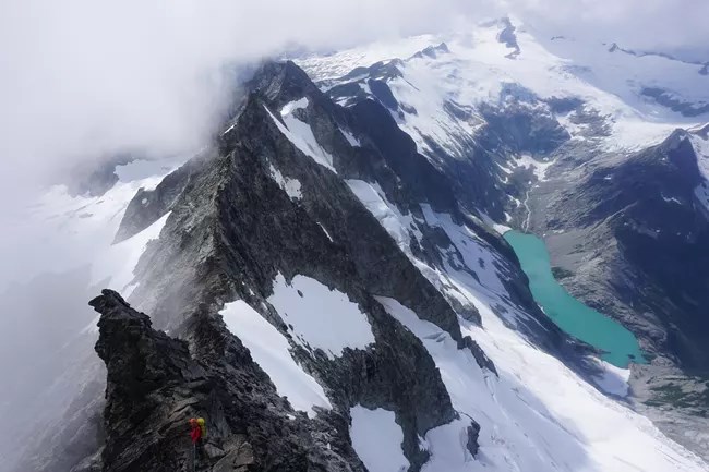 A ranger rappelling steep rocky ridge with glaciers and alpine lake in the background