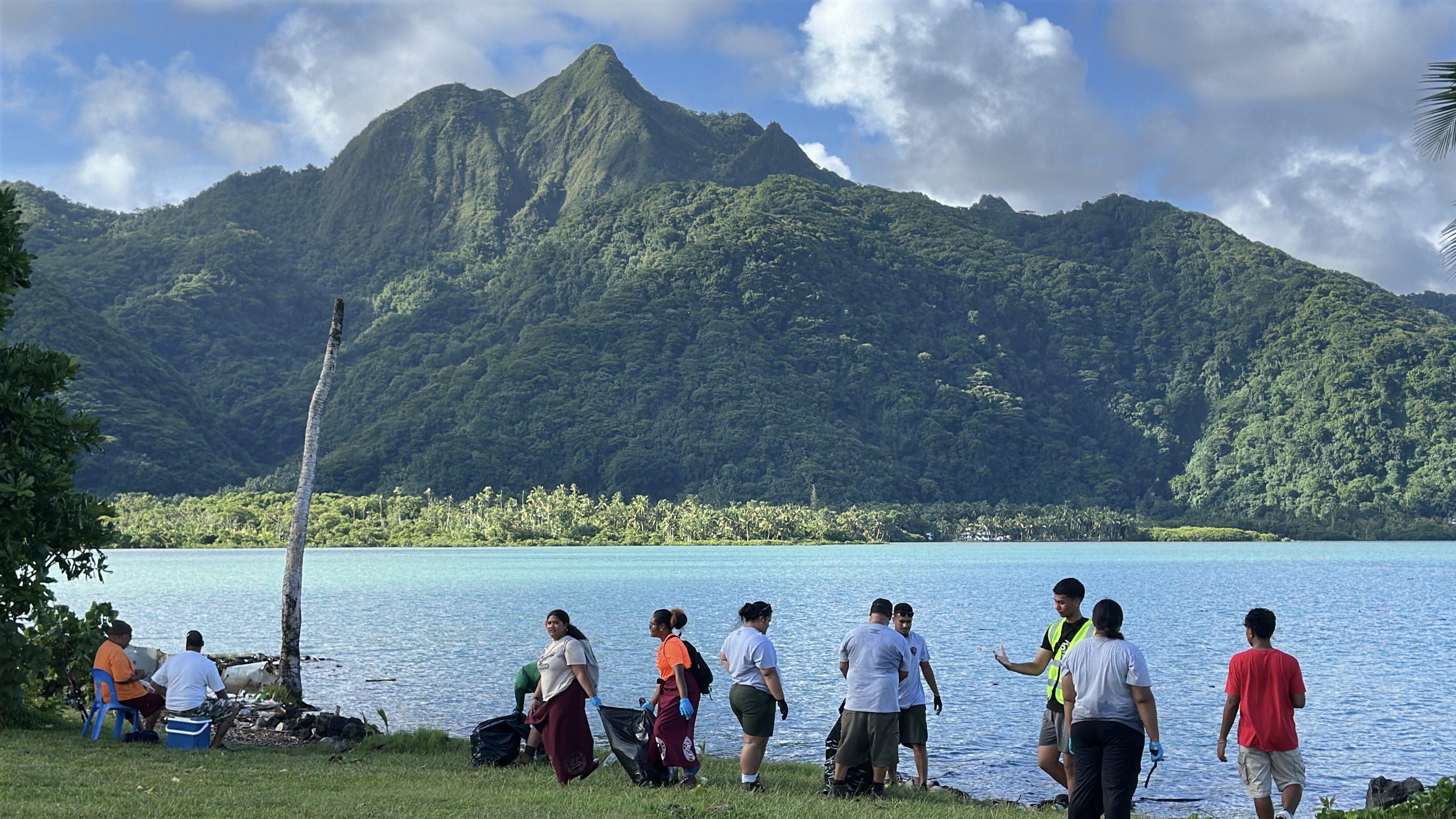Outdoors; Landscape view of young adults cleaning shoreline on green grass with blue water and green mountain in distance.