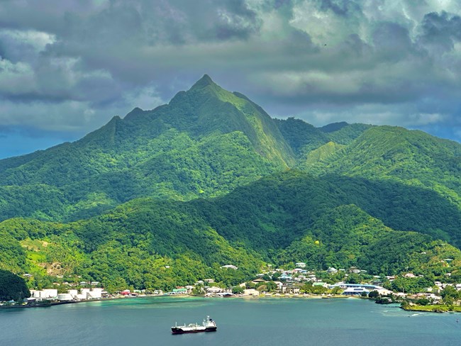Outdoors: Landscape view of boat and harbor with lush green mountain behind.