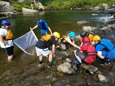 Several 6th graders and park ranger looking for aquatic macroinvertebrates in Clear Creek. They are all wearing lifejackets and whitewater helmets.