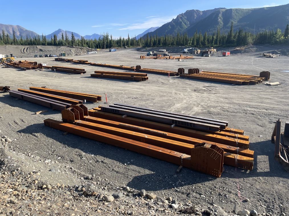 Steel beams organized on the ground in a gravel staging area.