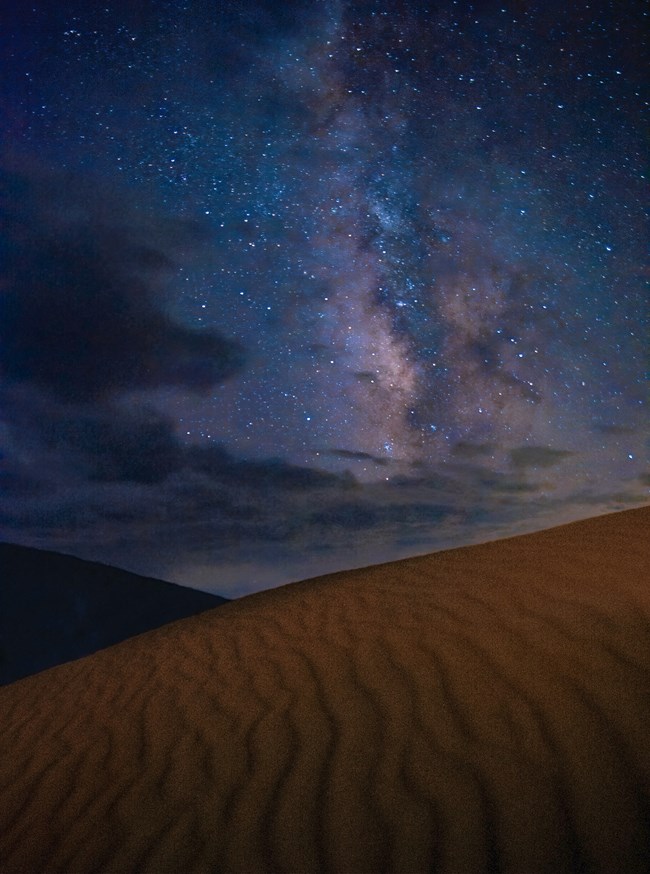 The Milky Way galaxy appears as a soft cloud with subtle colors in the starry sky over ripples in the sand.