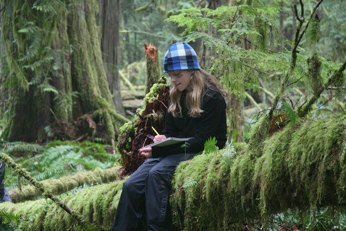 A girl writes in her journal while sitting on a log in a lush, green forest.