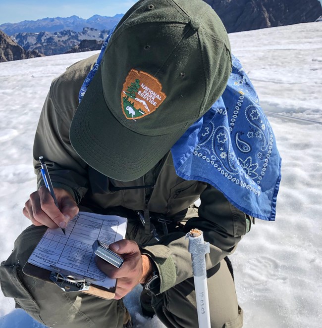 A person wearing an NPS ballcap crouches on a snow-covered glacier, writing on a datasheet.