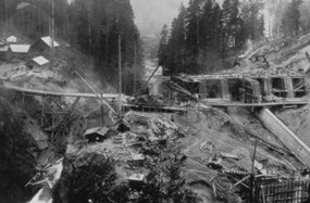 A historic, black and white photo of the Elwha Dam in the early stages of construction. An incomplete wooden frame is constructed over a canyon; water flows freely below. To the right, a concrete spillway is beginning to take form.