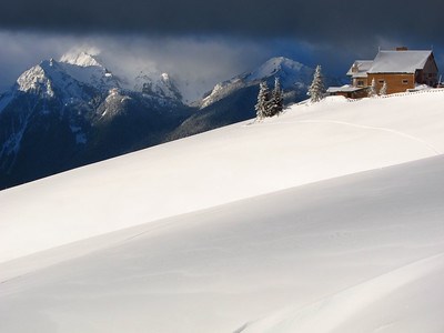 The Hurricane Ridge Visitor Center stands in contrast to the purely snow covered slope it stands on. In the background, a view of the Olympic Mountain Range rises through low-hanging, grey clouds.