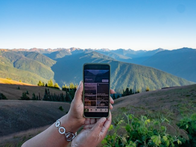 A hand holds a mobile phone with an outdoors app open on screen. A mountain range stretches beyond.