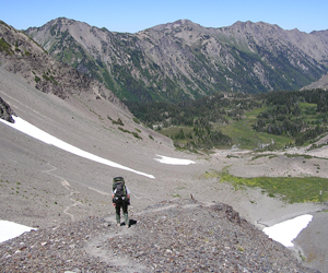 A hiker descends a steep, rocky trail in a mountain basin. This section of the basin is comprised of mostly scree - small, loose rock. Further down, the trail leads to a greener portion of the valley, with meadow growth and trees.