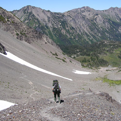 A hiker descends a steep, rocky trail in a mountain basin. This section of the basin is comprised of mostly scree - small, loose rock. Further down, the trail leads to a greener portion of the valley, with meadow growth and trees.