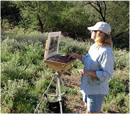 A painter stands in a brushy area while creating an image on canvass.