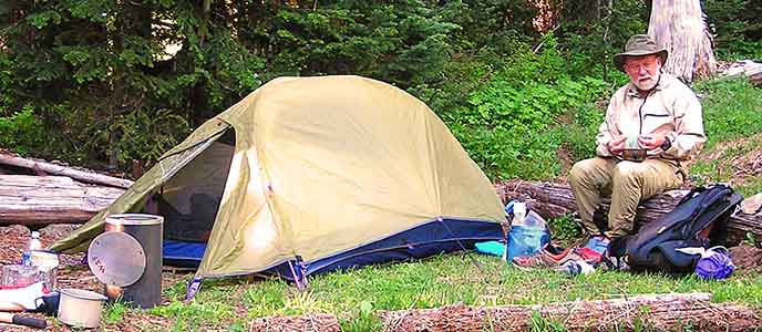Man in a wilderness campsite. He sits beside his tent with lots of backpacking gear nearby.