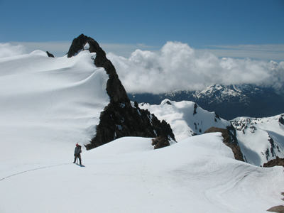 A mountaineer stands on a snowy slope high in the mountains, with lower snowcapped mountains lying below in the background.