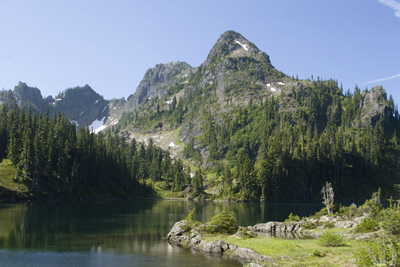A rocky mountain peak juts up behind a subalpine lake. The shores of the lake are comprised of a mix of forest and meadow.