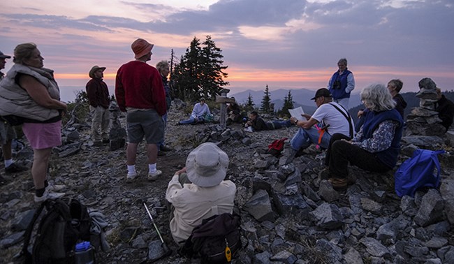 A group of people seated and standing on rocky terrain during sunset, participating in an outdoor educational program at Oregon Caves National Monument. The group is surrounded by trees and distant mountain views as they engage in a discussion.