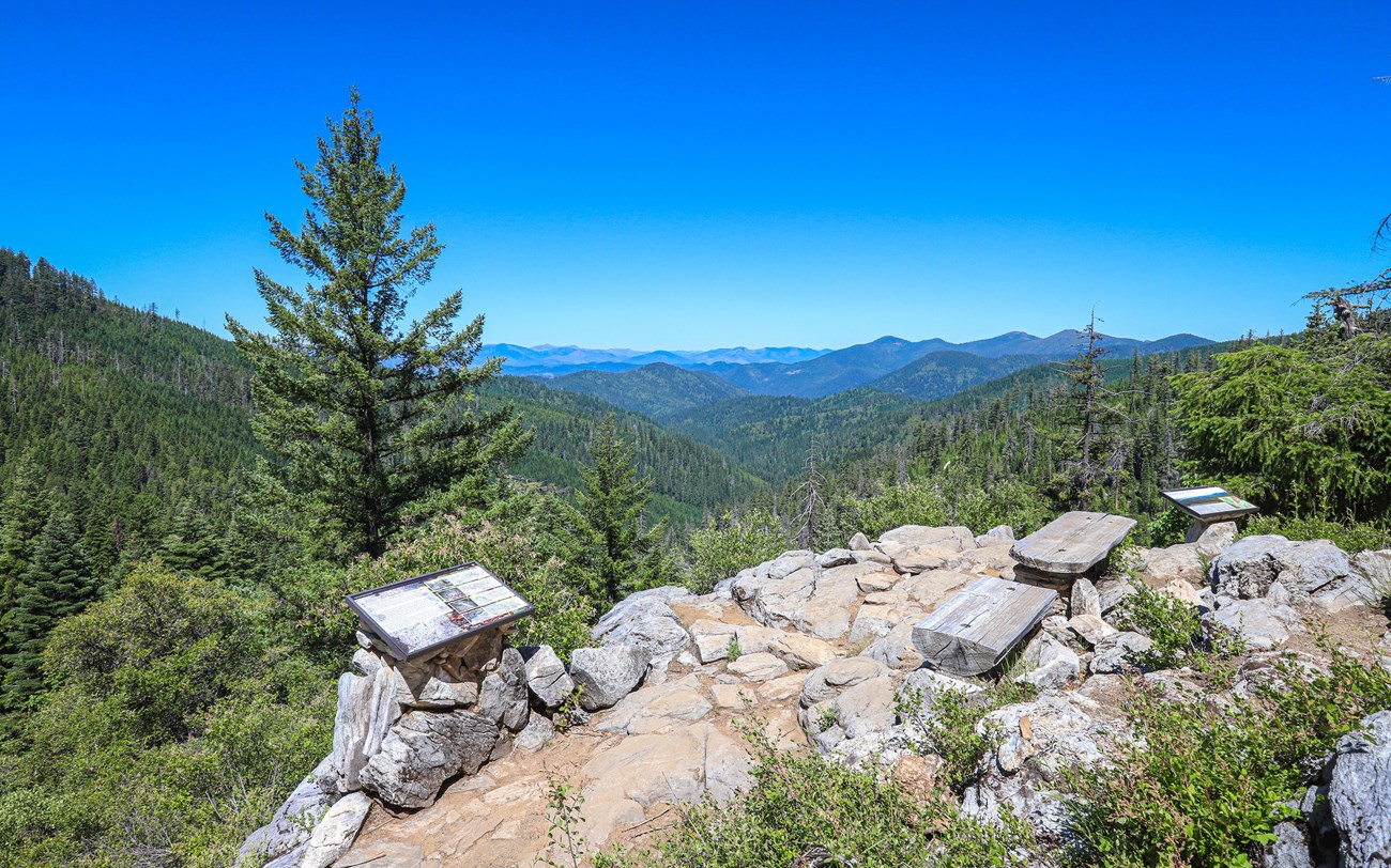 A panoramic view from the Cliff Nature Trail at Oregon Caves National Monument, showing a rocky overlook with interpretive signs and benches surrounded by lush green forests and distant mountain ranges under a clear blue sky.