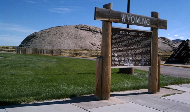 A large wooden sign engraved with the word "Wyoming" and a large paragraph of text stands in front of a large rounded rock buttress with green grass.