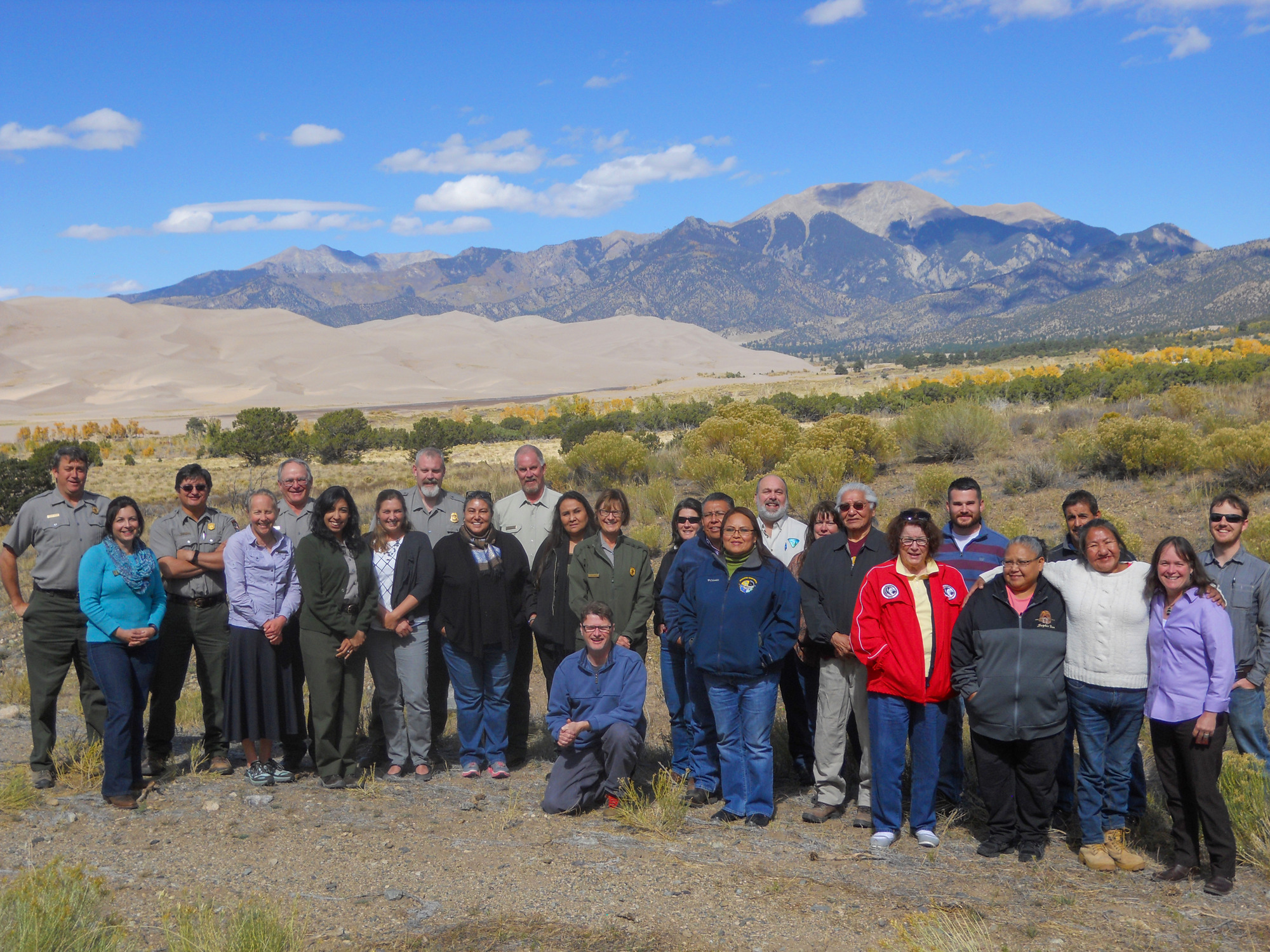 Representatives from the Southern Ute, Ute Tribe of Uinta and Ouray, Ute Mountain Ute, Jicarilla Apache, and Navajo tribes stand with Interior Department staff after a tribal consultation. In the background are the dunes and Sangre de Cristo Mountains.