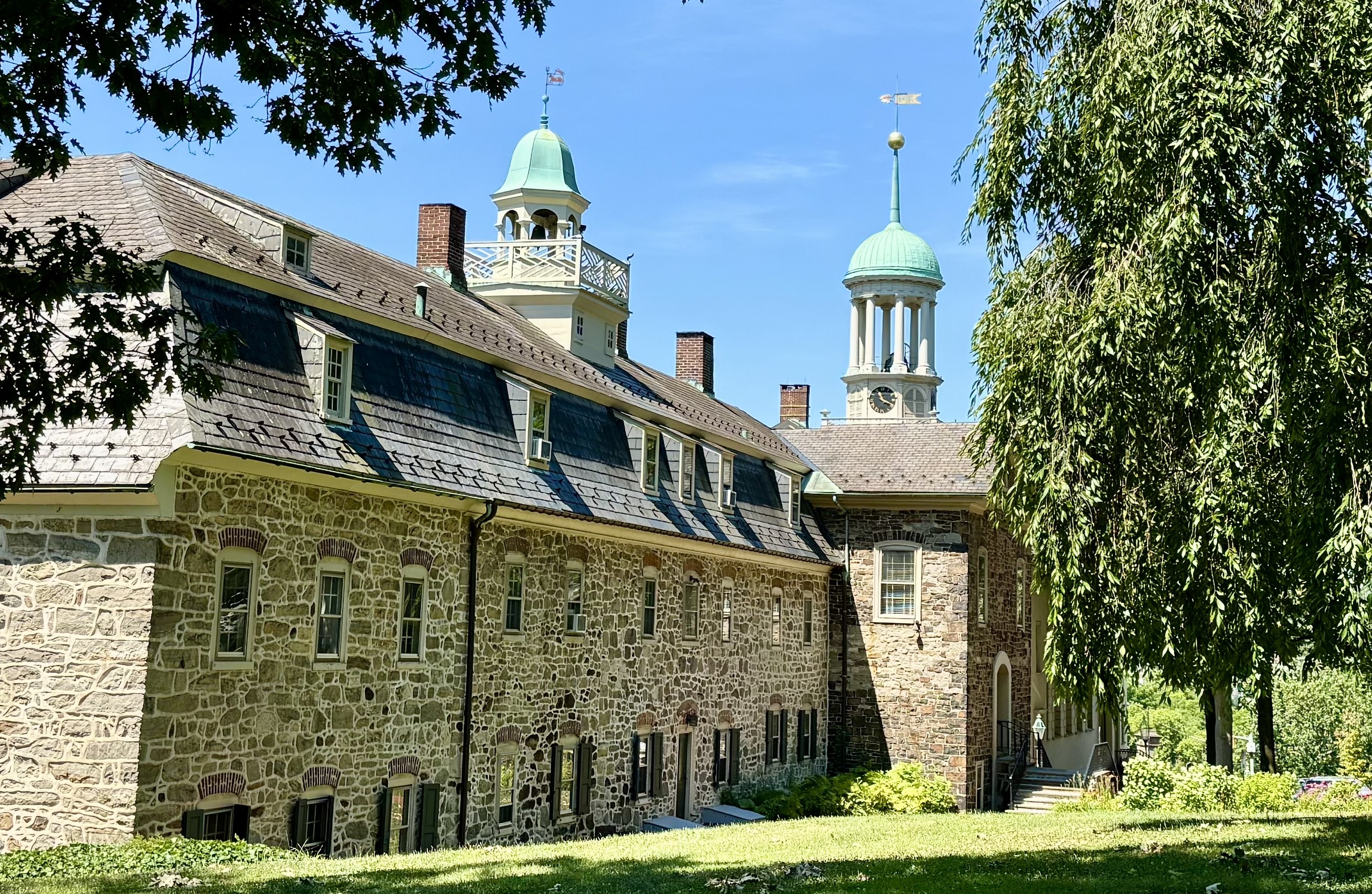 To the left, a stone building stands with multiple windows and a belfry; on the right is a large tree.