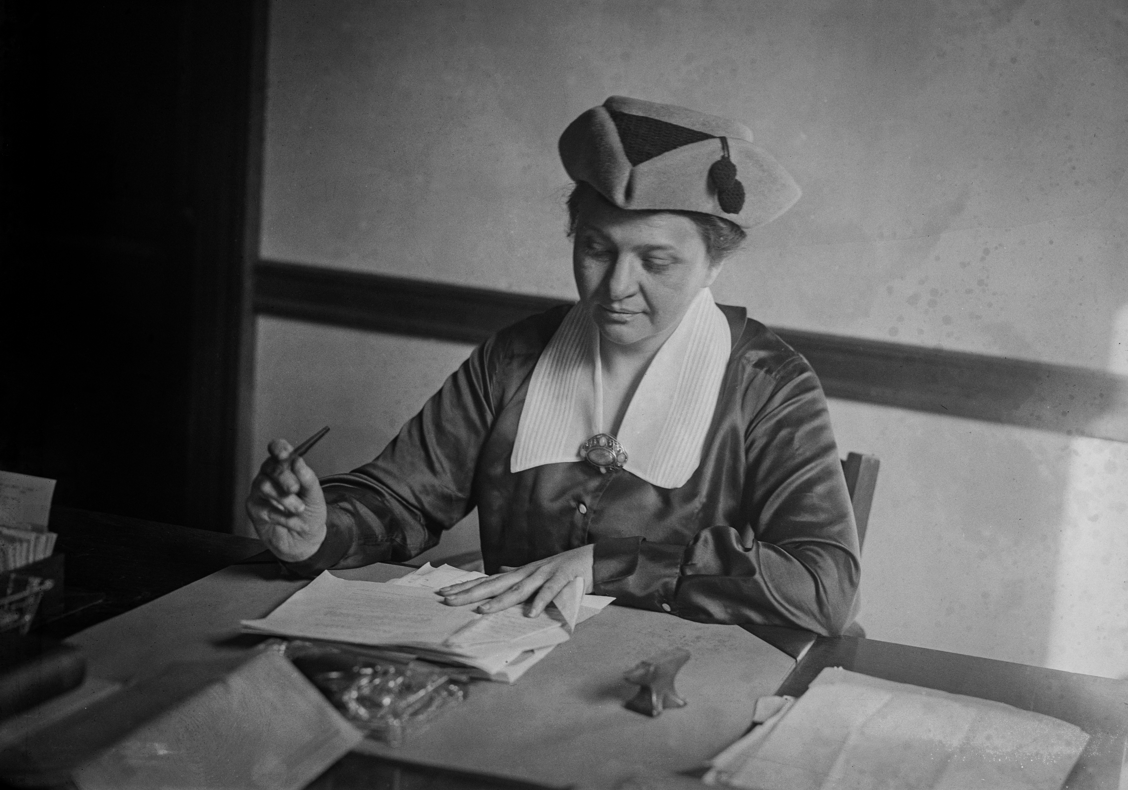 In black and white, a woman in a triangular hat and a satin top with a large white collar hols a pen over a stack of papers on a desk