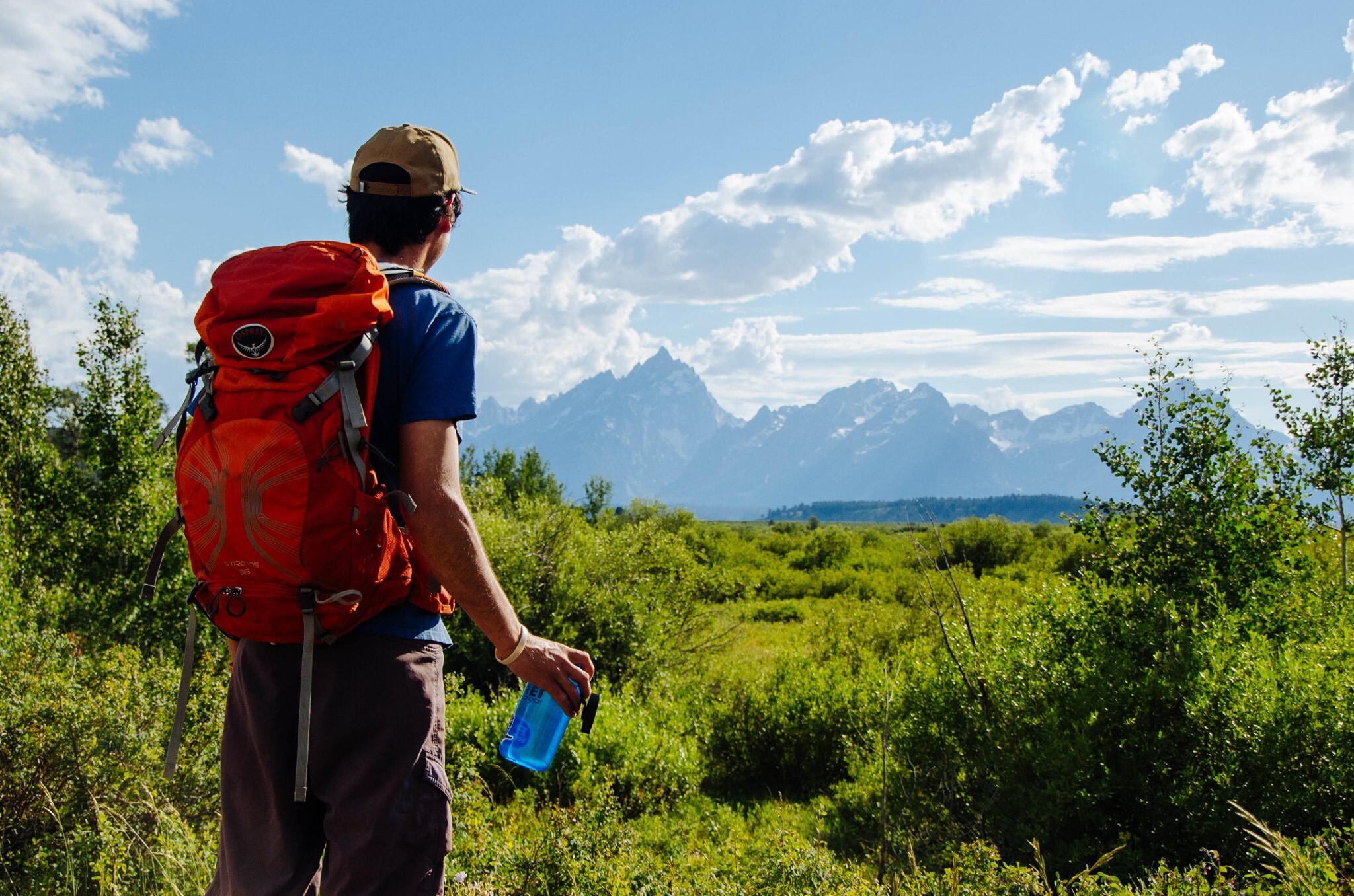 A hiker looks out on Teton Range holding a water bottle and wearing a backpack.