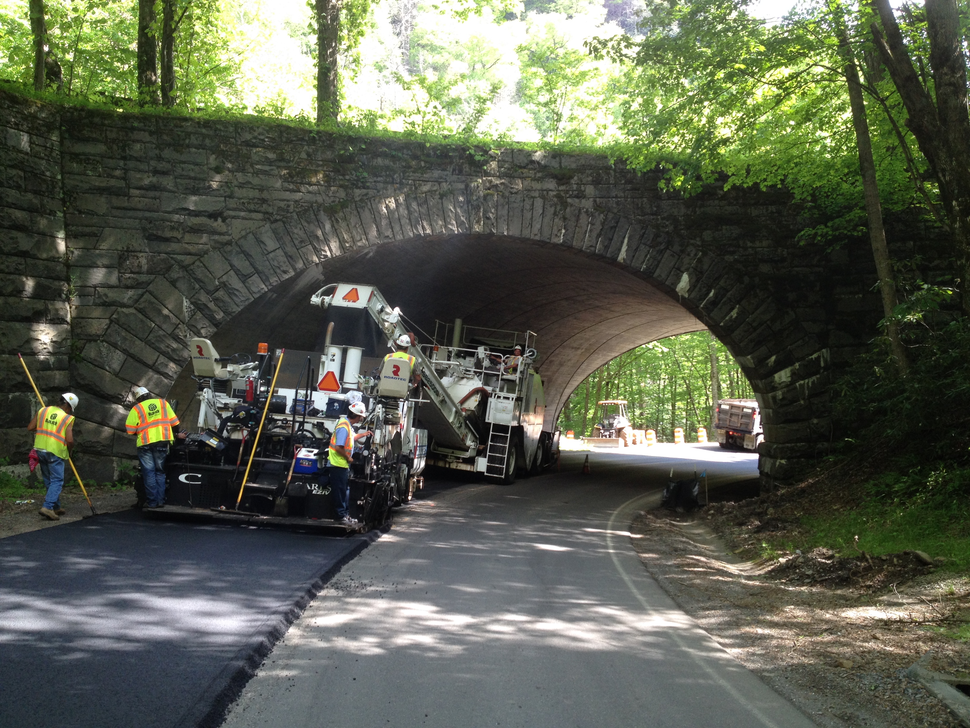 Road crew paves a road just before a stone bridge overpass.
