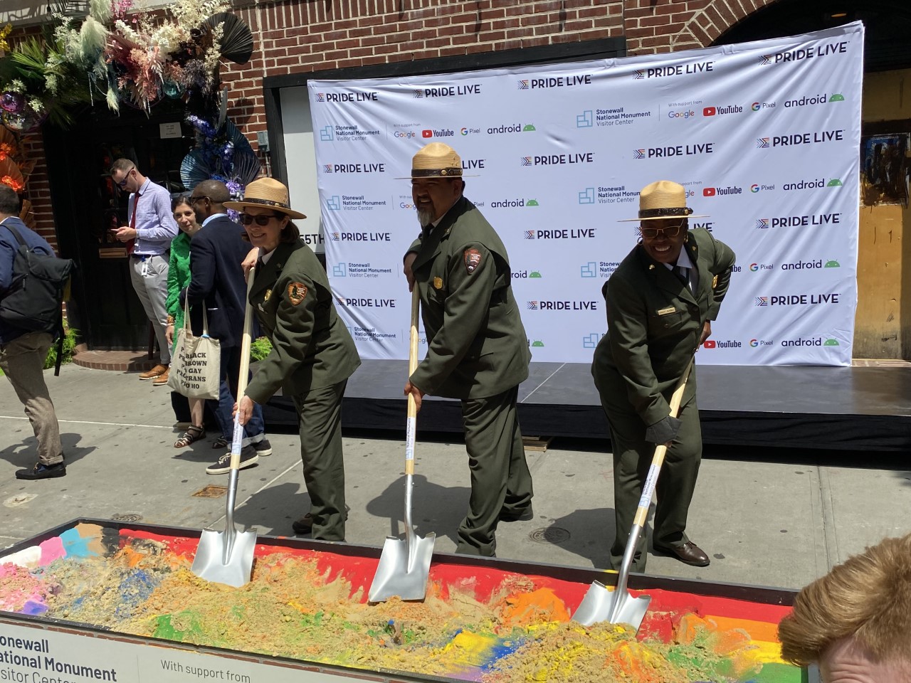 Three park rangers in green uniforms plant shovels into rainbow-colored dirt in front of a backdrop reading "Pride Live".