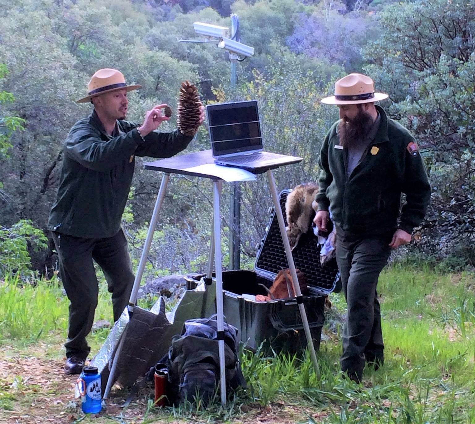 Ranger in front of monitor holding a small pinecone and a large pinecone with another ranger nearby.