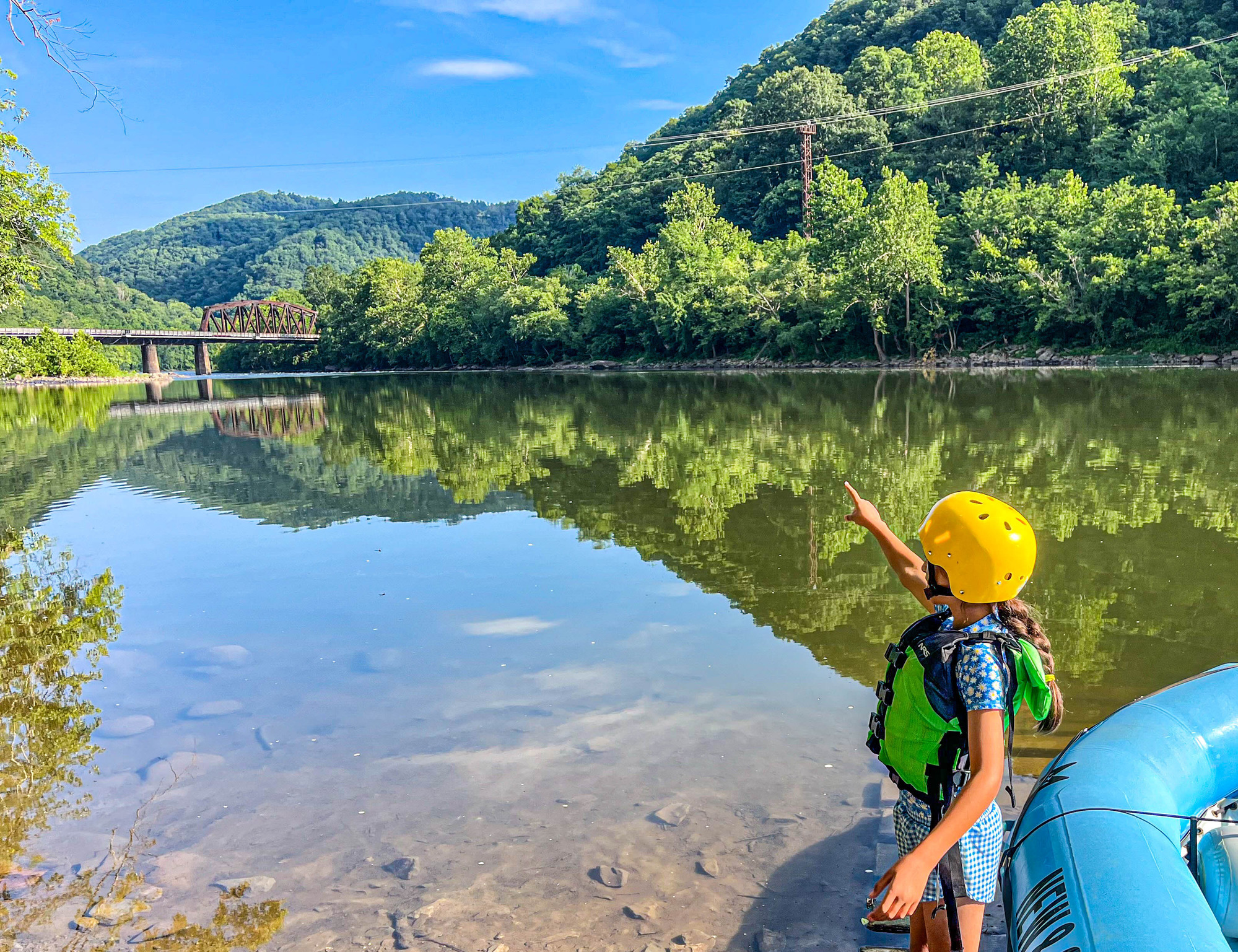 Kid standing next to a raft pointing to a bridge over a river lined with trees