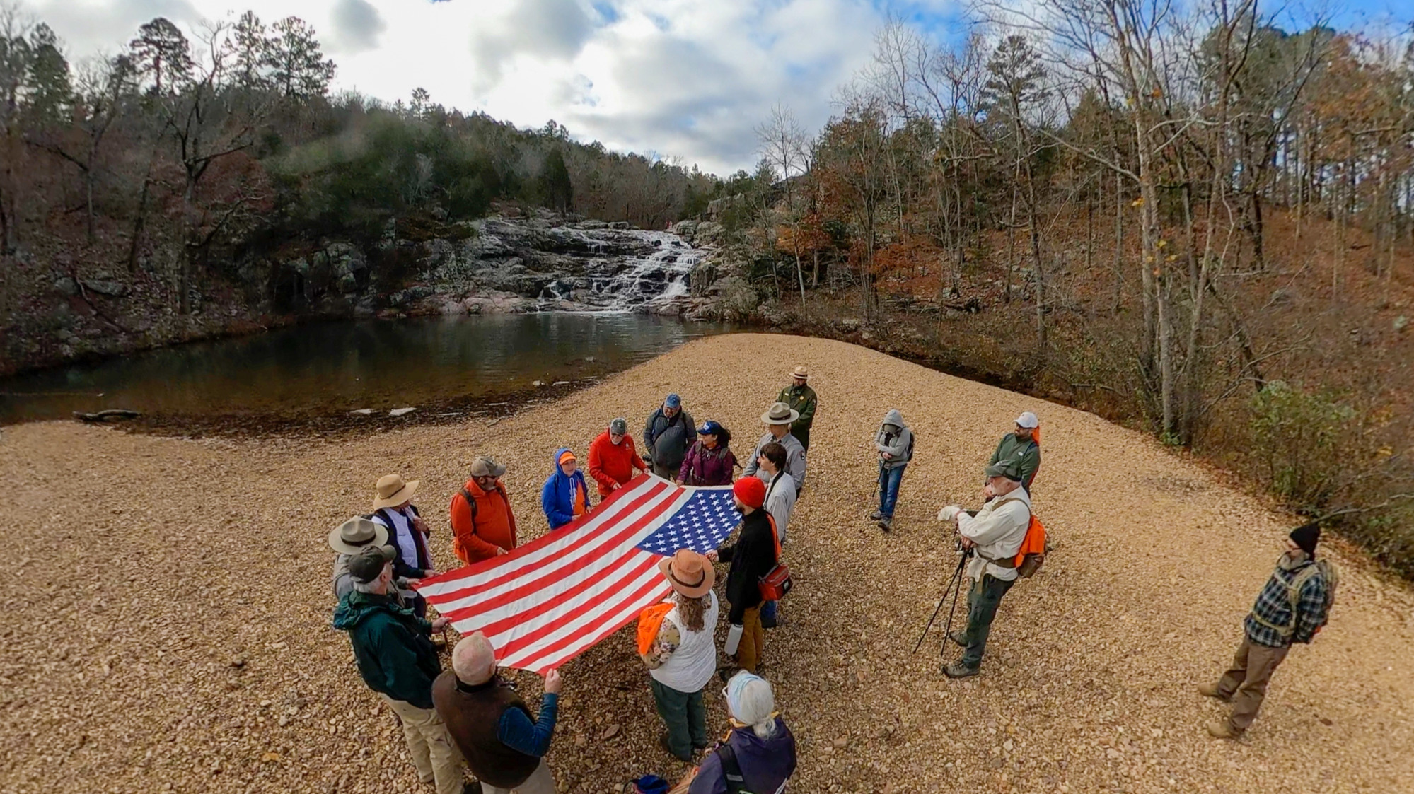 A group of people dressed in hiking clothes standing around and holding an unfolded American flag wtih a waterfall, river, and trees without leaves in the background.