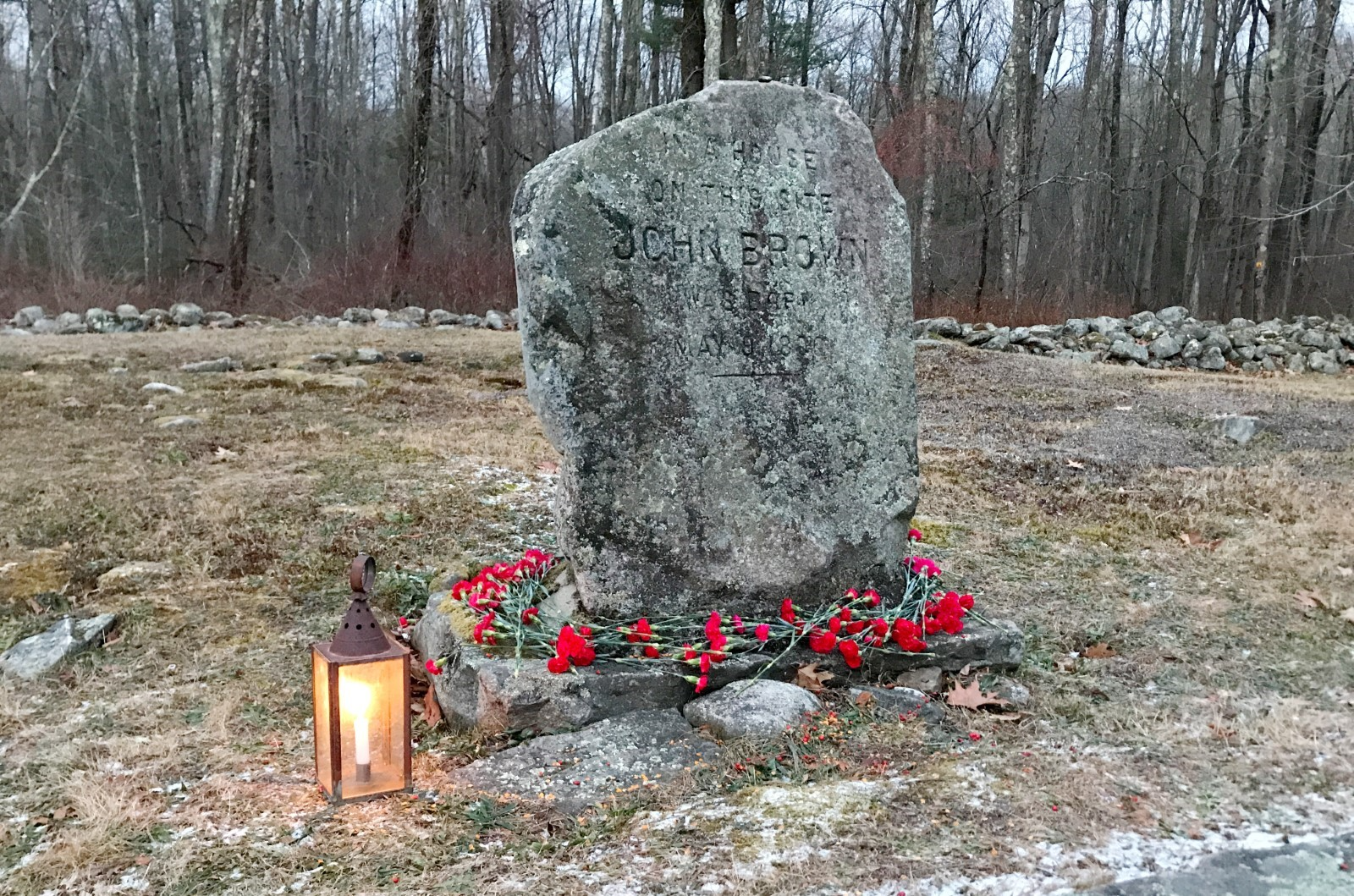 A heavily aged and stone marker with "John Brown" carved into it. The base is surrounded by red flowers and there is a lit lantern to its left.