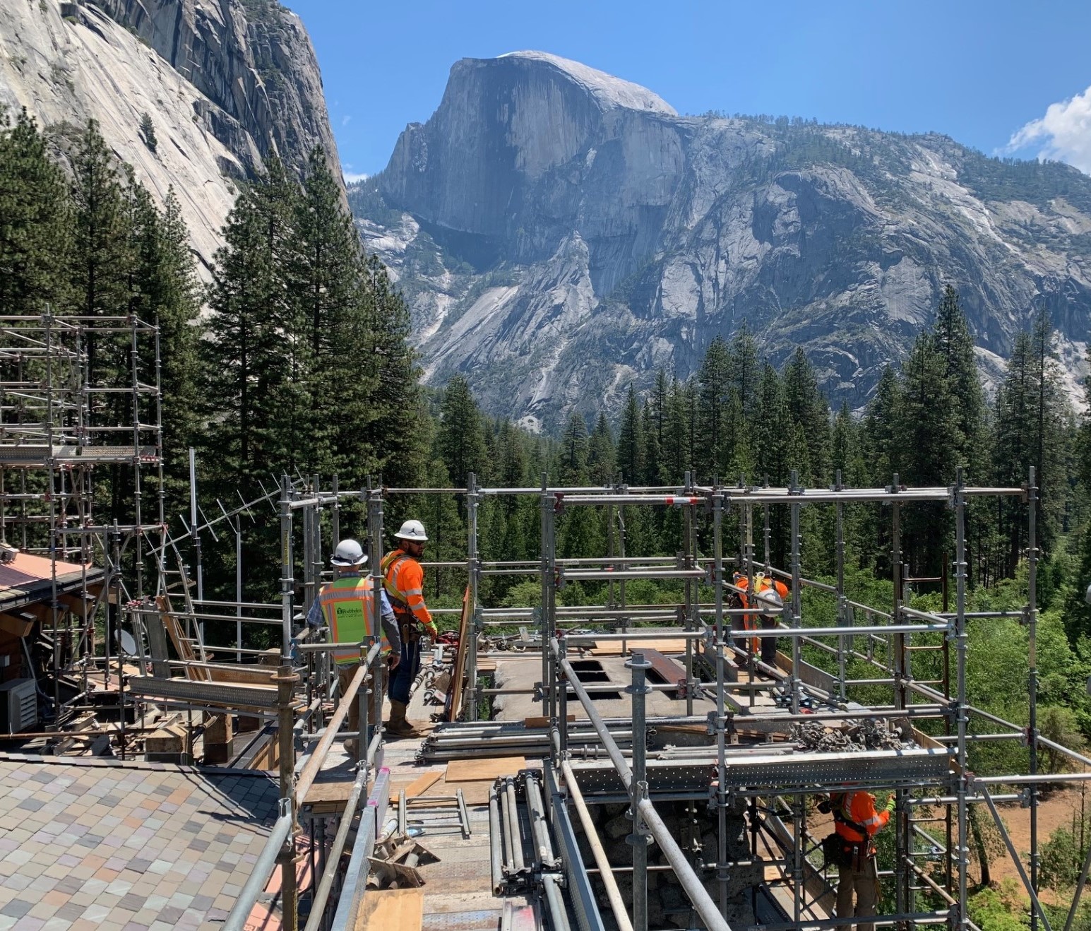 Two construction workers in orange and yellow safety vests stand on scaffolding in front of a large, sheer granite mountain.