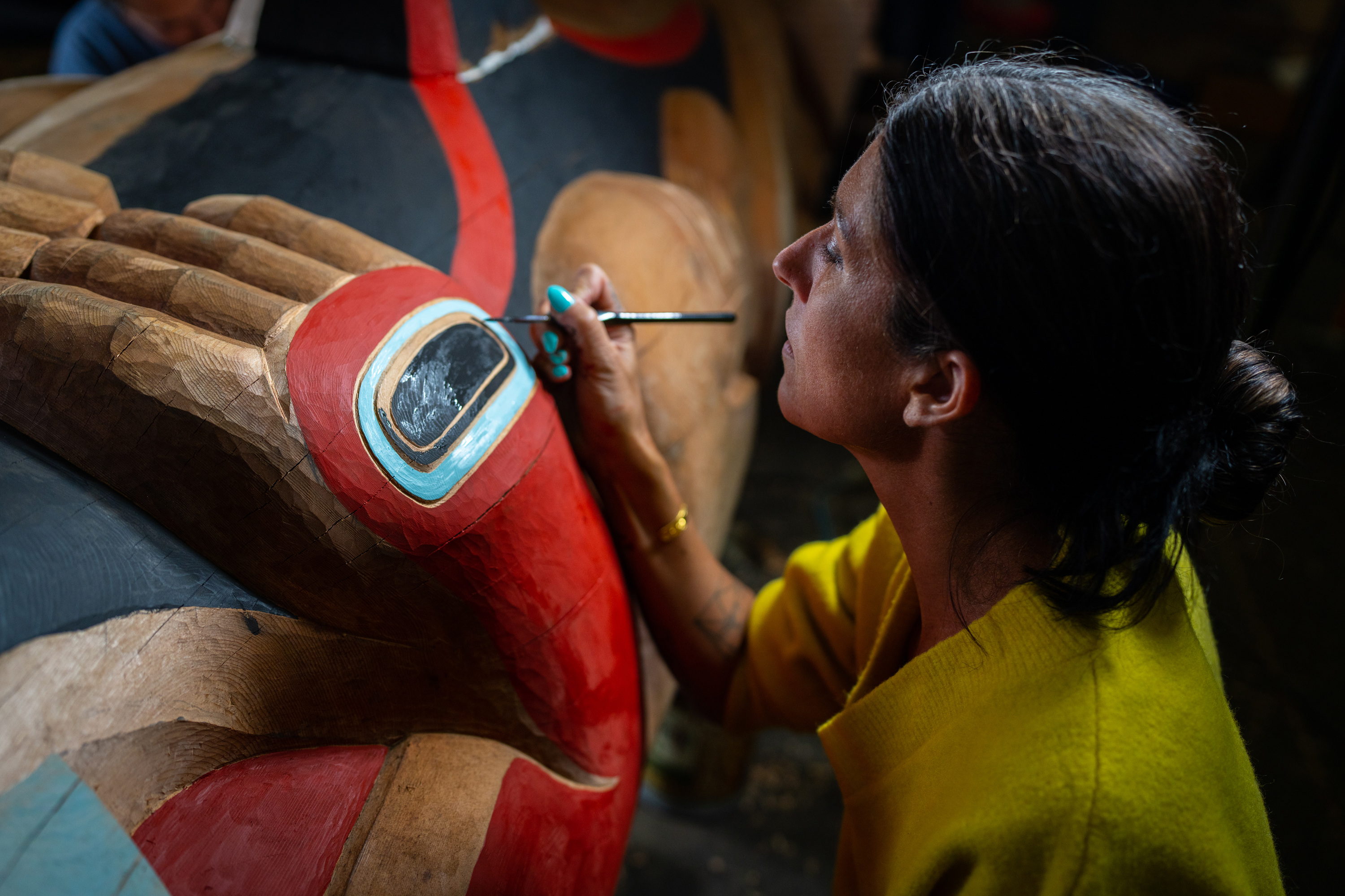 An artist paints a Kaagwaantaan totem pole multiple colors, including red, blue, and black.
