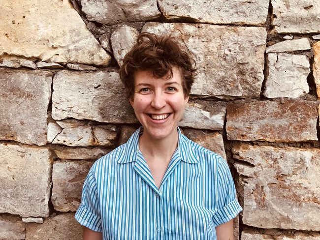 A person with brown hair and a blue striped shirt standing in front of a stone wall.