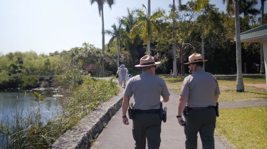 Two rangers walking down a path next to a body of water with members of public ahead of them in the distance.