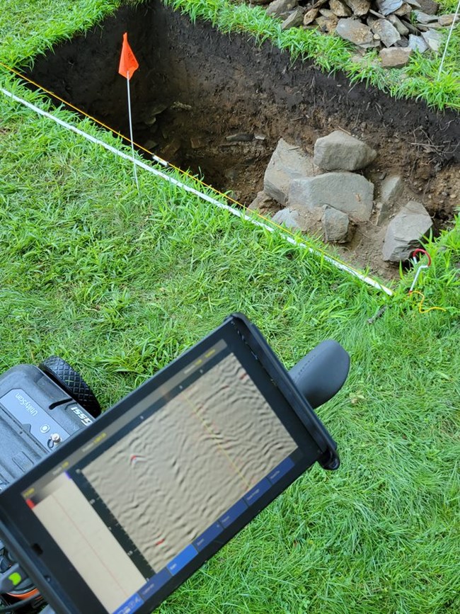 A GPR machine with a scanner sits next to an open excavation unit with rocks peeking out
