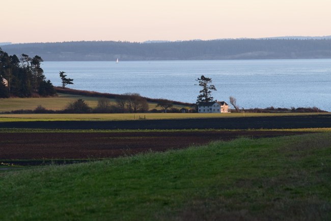 An old house sits between rich farmland and the sea.  A sailboat can be seen in the distance.