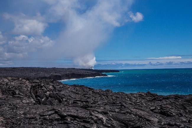 Black volcanic rock along the blue coastal waters and volcanic steam rising in the background.