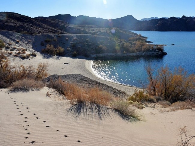 A trail of footprints on a sandy beach by a blue lake and rocky hills in the background.