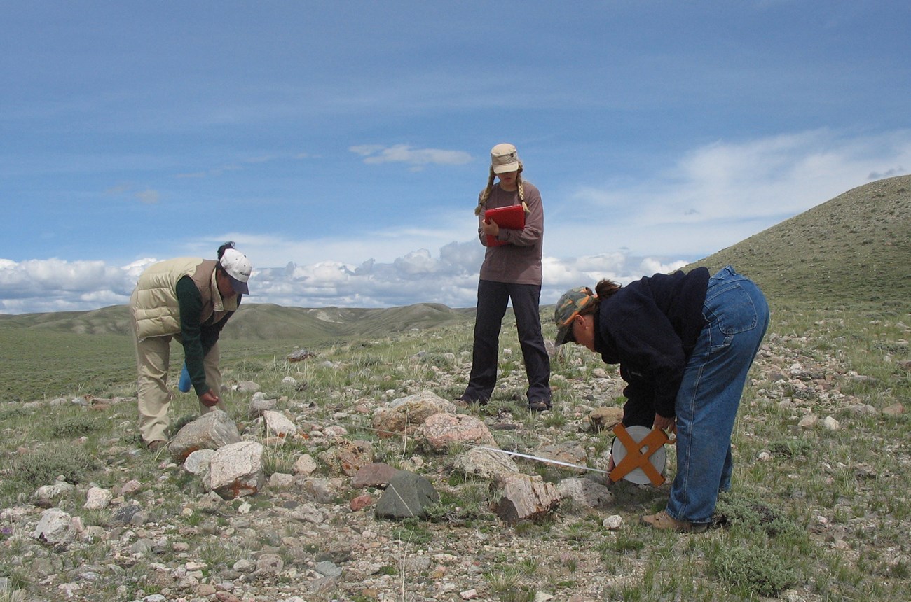 Two students measure a rock pile located in sagebrush and grass covered mountains, while a third student records the data.