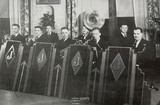 A black and whit image of men in suits standing behind chairs.