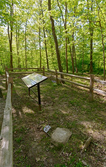 Wood fence in a forest surrounding a wayside sign and a gravesite marked with stone markers, under a canopy of green leaves