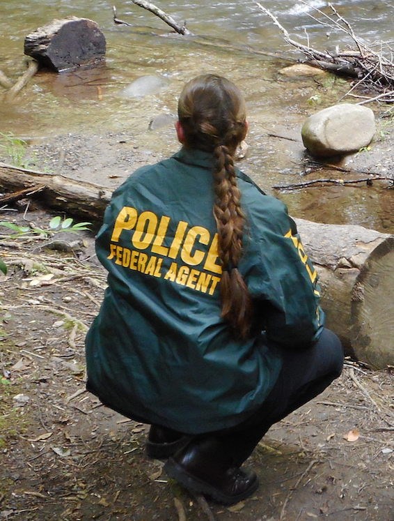 An ISB Special Agent crouches near a stream bank. She wears a green jacket with the words "Police" and "Federal Agent" printed across the back.