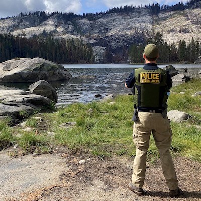 An ISB Special Agent stands along the shore of a lake and looks out across the water to the distant forested cliff.