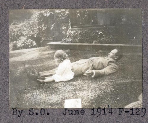 A child sits beside a man in suit who is laying on his back in the grass, 1914