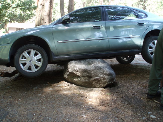 A car high-centered on a large rock, with its wheels lifted off the ground and the undercarriage resting on the rock