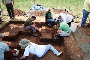Six people, all seated or kneeling, carefully dig in a square excavation block surrounded by tools and tarps. Two other individuals stand at the edge of the square block.